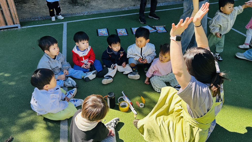 꼬꼬마어린이집 교사와 영아들이 바깥에 둘러앉아 함께 노는 상호작용 모습